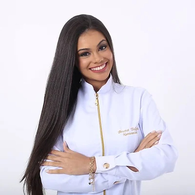 Bruna Talyta de jaleco branco bordado com seu nome, sorrindo com os braços cruzados